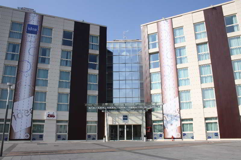 Idea Hotel Milano Malpensa Airport Case Nuove  Esterno foto The photo shows a modern hotel building with a prominent glass entrance. The façade features a mix of materials, including light-colored stone and dark panels. Large banners or displays adorn the sides of the building, adding visual interest. The are