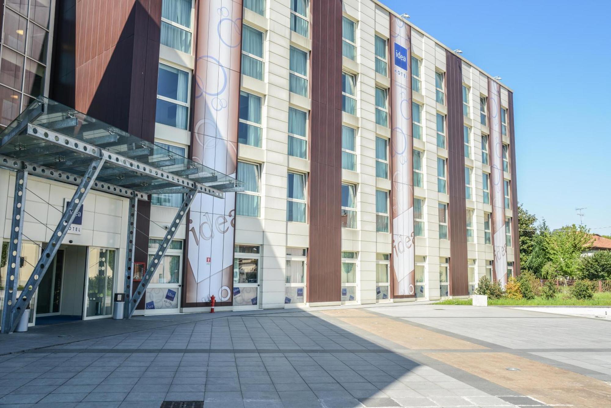 Idea Hotel Milano Malpensa Airport Case Nuove  Esterno foto The photo shows the exterior of a modern building, likely a hotel or office complex. The structure features a combination of glass and metal elements along with a concrete facade. There are large windows, and vertical stripes of wood-like material ad