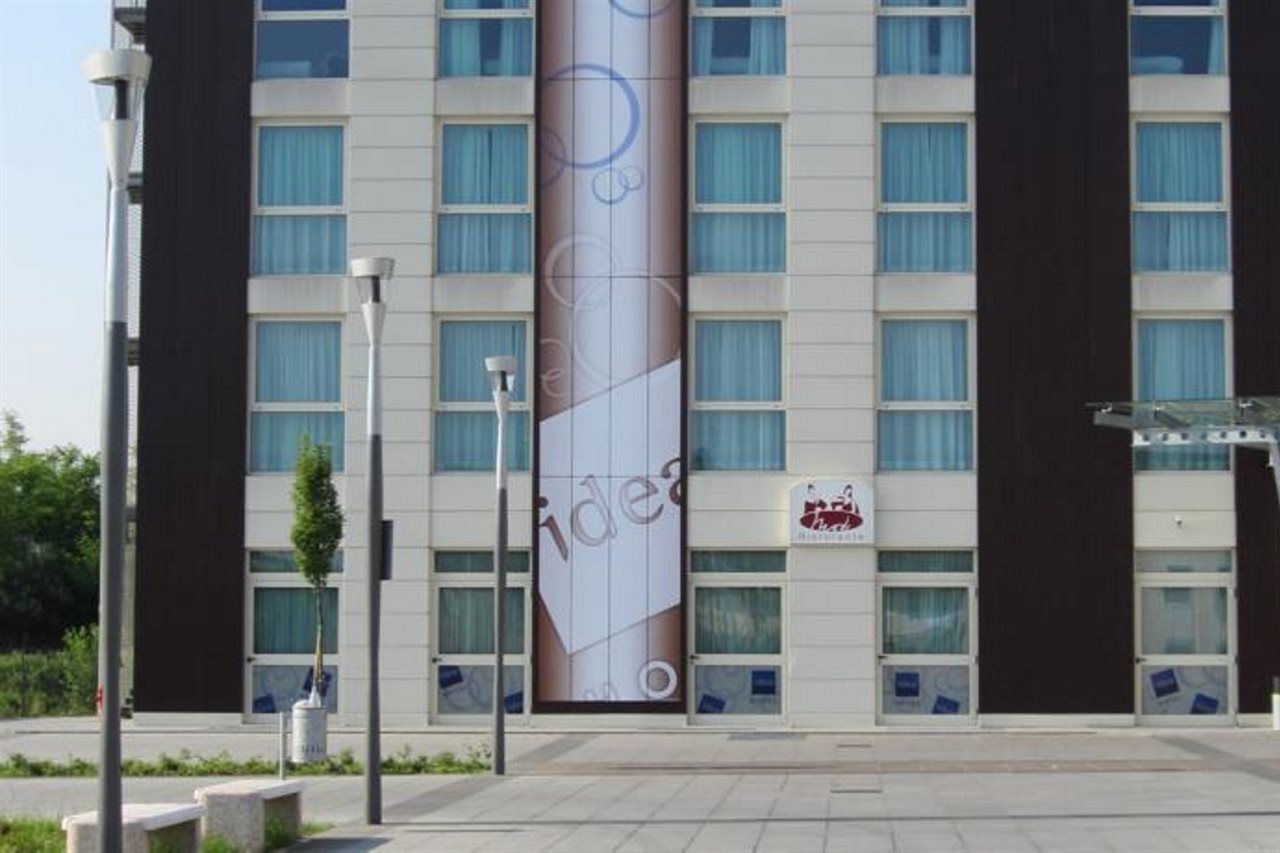 Idea Hotel Milano Malpensa Airport Case Nuove  Esterno foto The photo shows a modern building facade. The exterior has a mix of white and dark panels, with several large windows featuring blue curtains. A prominent banner or advertisement runs vertically down the center of the building, showcasing an abstract