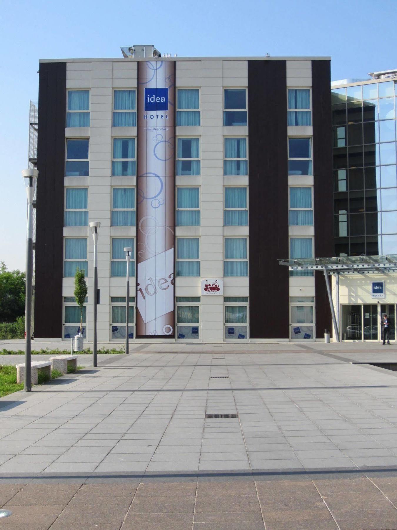 Idea Hotel Milano Malpensa Airport Case Nuove  Esterno foto The photo shows the facade of a multi-story building, likely a hotel. It features a combination of light and dark exterior panels. In the center, there is a large banner with a design, possibly advertising or displaying a logo. The entrance appears t