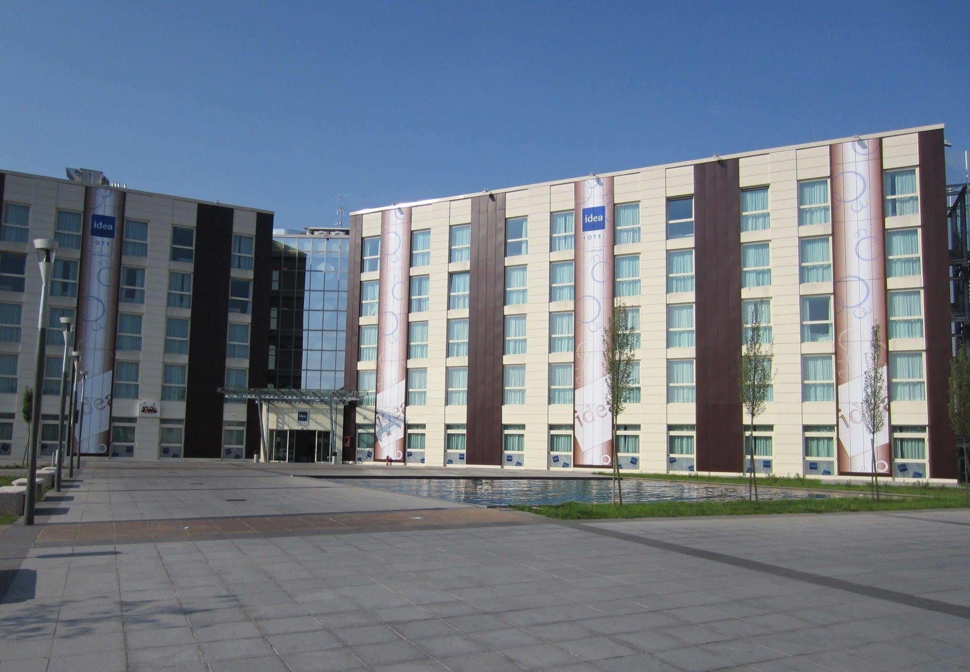 Idea Hotel Milano Malpensa Airport Case Nuove  Esterno foto The photo shows a modern multi-story building, likely a hotel or office complex. The building features a combination of light-colored and dark-colored panels, with large windows allowing for natural light. There are promotional banners displayed on t