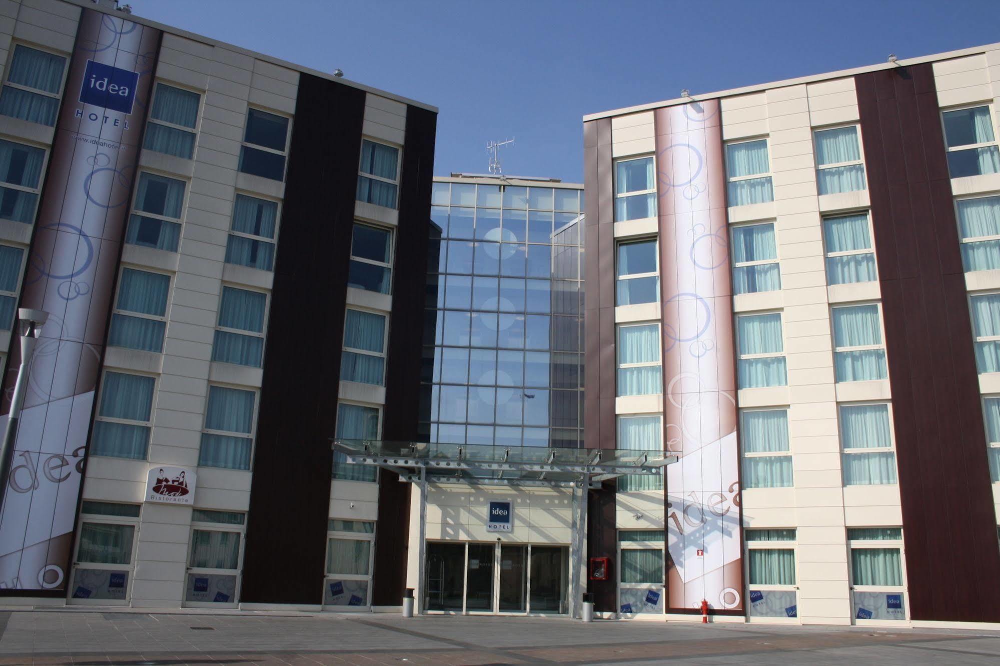 Idea Hotel Milano Malpensa Airport Case Nuove  Esterno foto The photo shows the exterior of a modern hotel building. It features a large glass entrance surrounded by two taller sections composed of light-colored and dark-colored panels. The left side has a banner displaying the hotel name, and there are large