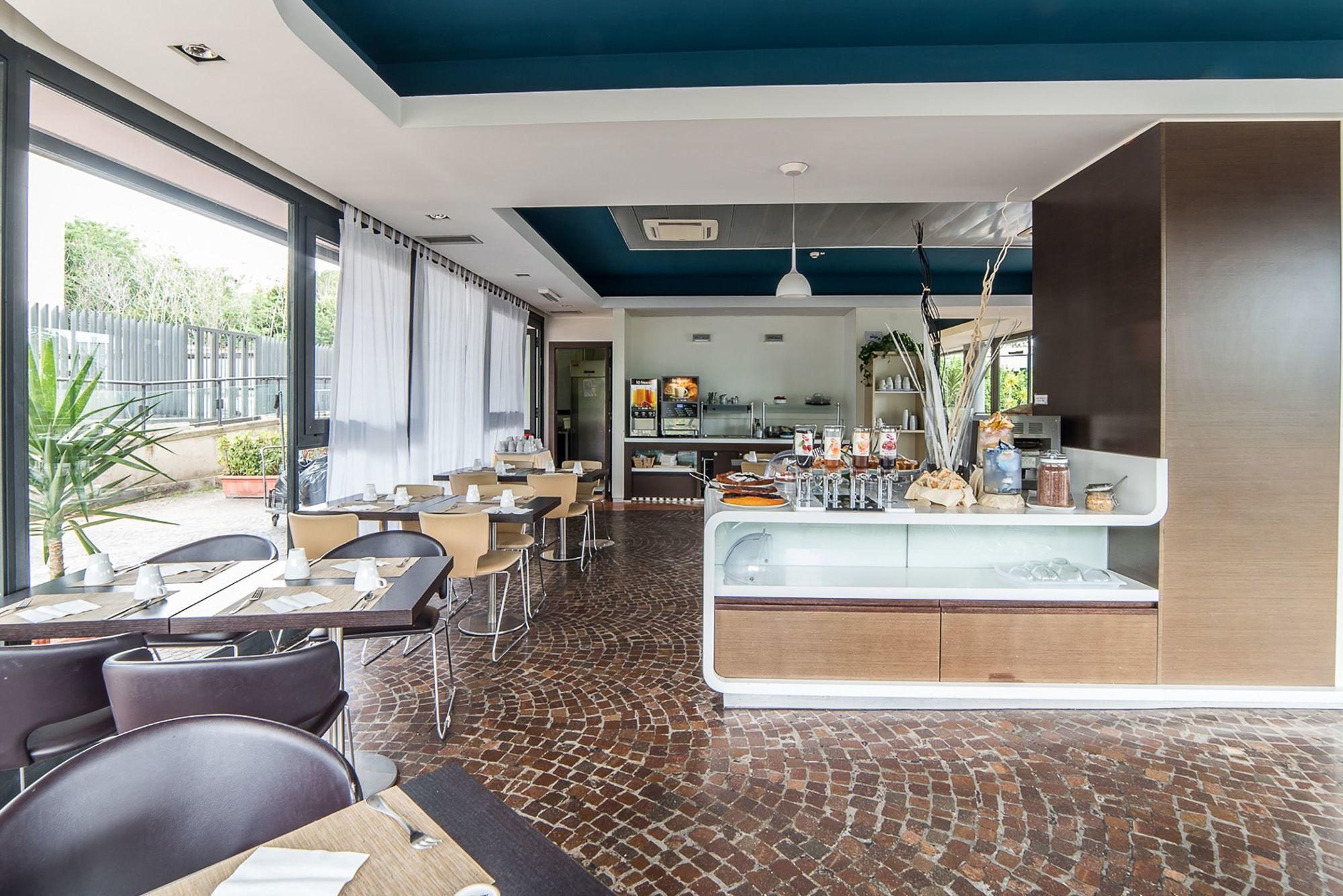 Idea Hotel Milano Malpensa Airport Case Nuove  Esterno foto The photo shows a modern dining area, likely part of a restaurant or cafe. The interior features large windows that let in natural light and offer a view of greenery outside. The decor includes a mix of wooden elements and colorful accents, with a bl